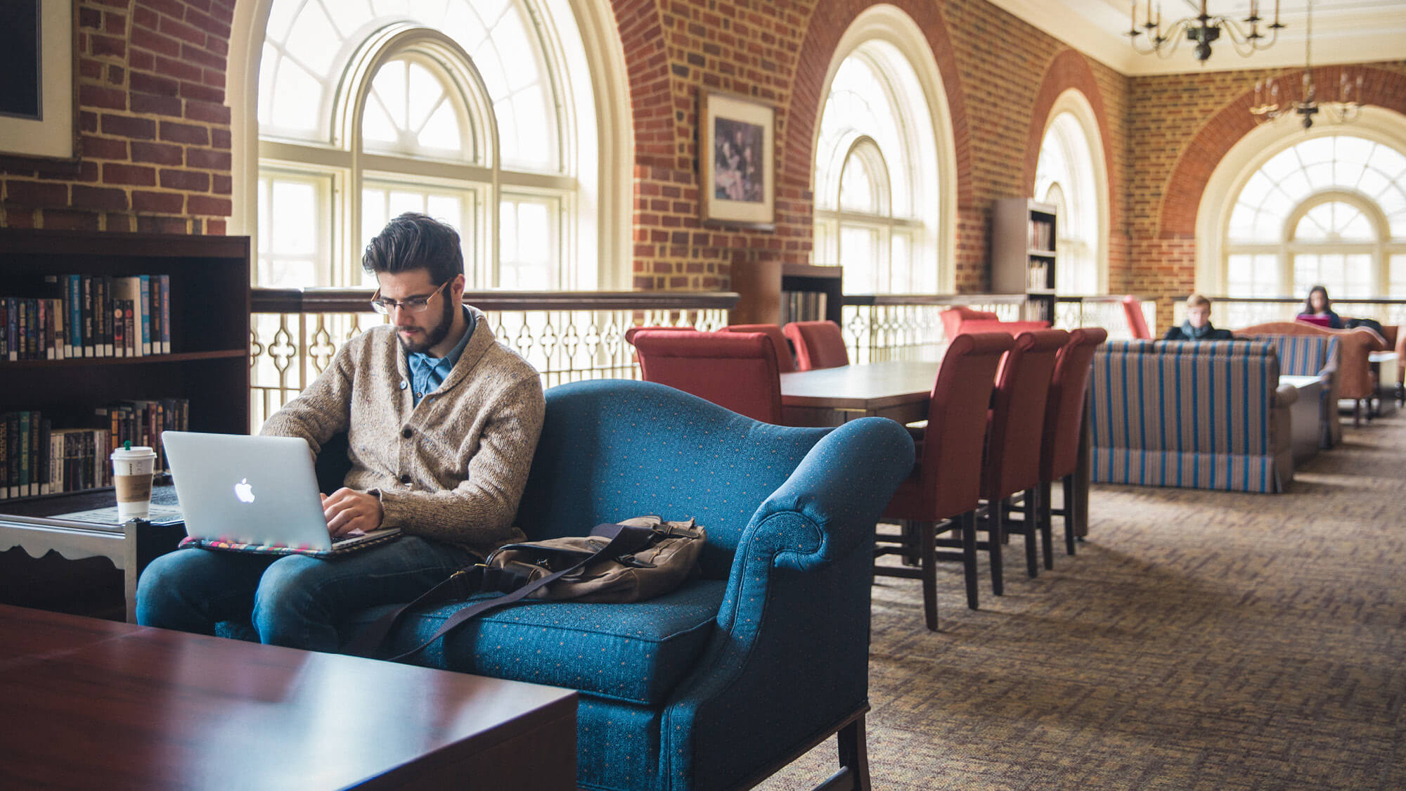 A student sitting on a blue couch in the Regent University Law Library, working on a laptop in a study area with arched windows and natural light.